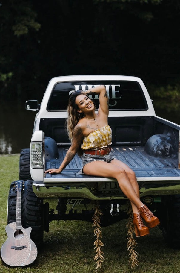 Woman wearing Culture Girl yellow palaka print  bandeau sitting on a pickup truck bed with a guitar leaning against it, outdoors.