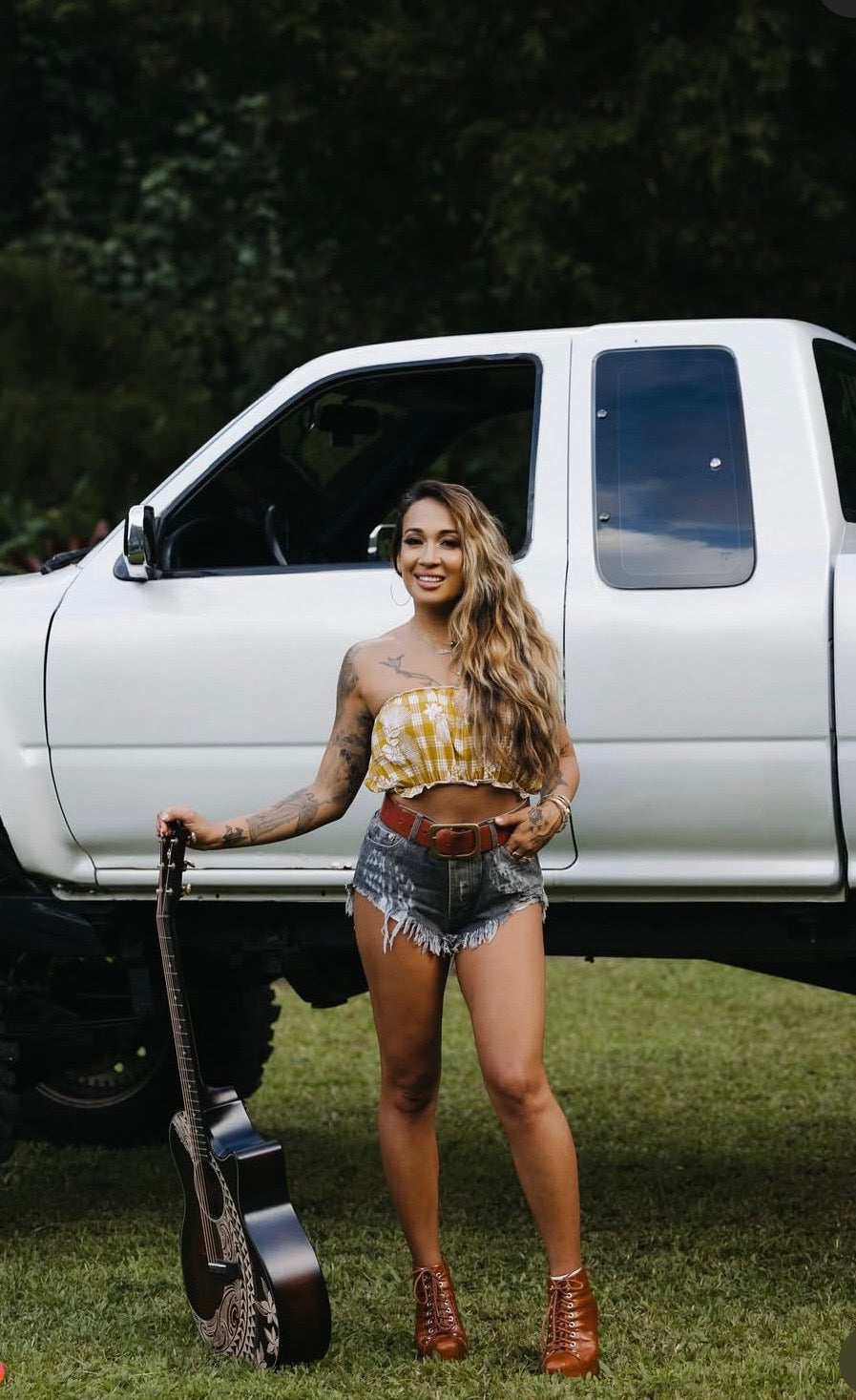 Woman standing in front of a white truck holding a guitar, wearing a Culture Girl yellow palaka print top and denim shorts.