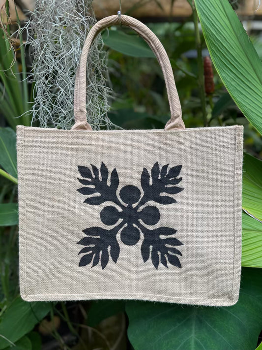 Beige tote bag with black ulu design hanging among green leaves