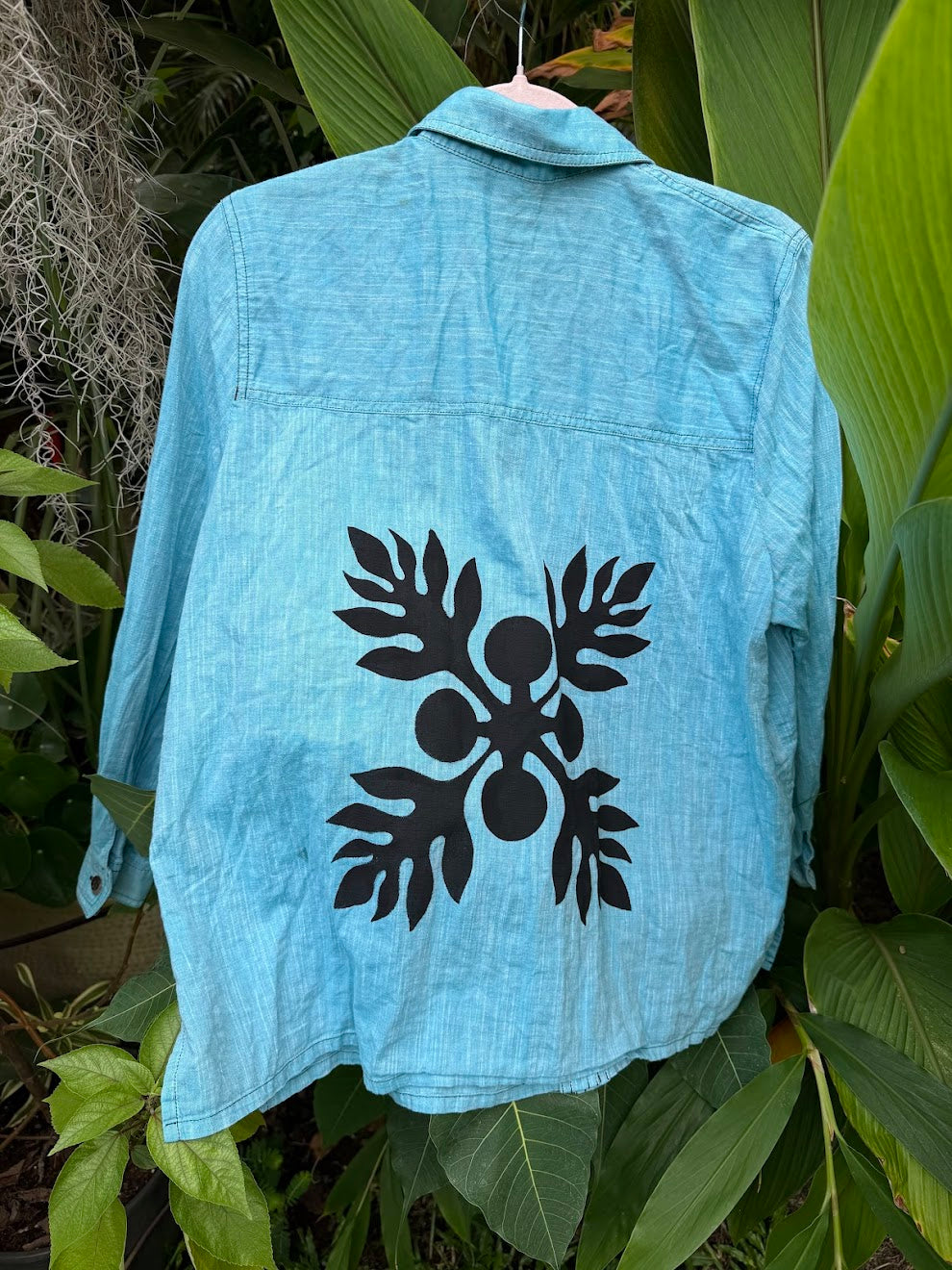 Light blue denim button down jacket with a black Hawaiian Ulu print design on a green leafy background